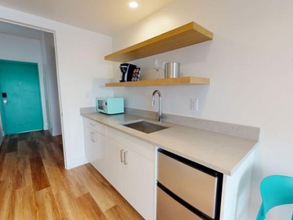 This image depicts a small kitchenette with a sink, a mini fridge, open shelves, a microwave, and a teal chair, leading to a room with a turquoise door.