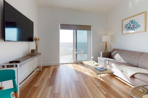 A modern living room with a mounted TV, sofa, coffee table, and sliding glass doors opening to a sea view. A framed picture and lamp are present.