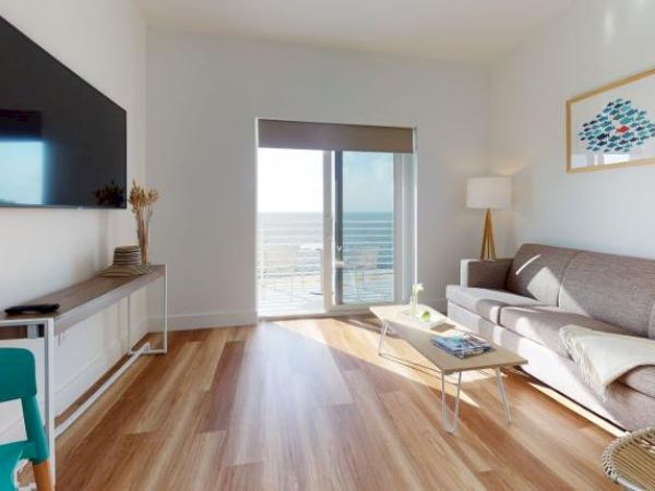 A modern living room with a mounted TV, sofa, coffee table, and sliding glass doors opening to a sea view. A framed picture and lamp are present.