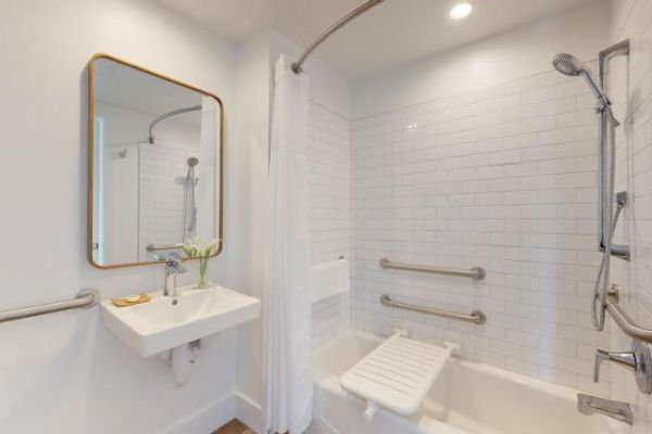 This image shows a bathroom with a white tiled shower area featuring grab bars, a fold-down seat, a wall-mounted sink, and a mirror.