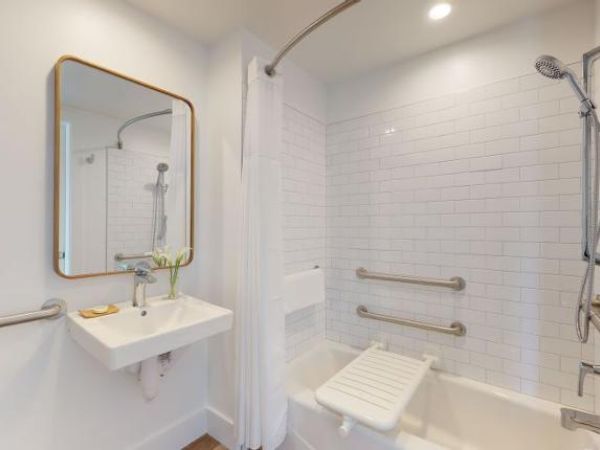 This image shows a bathroom with a white tiled shower area featuring grab bars, a fold-down seat, a wall-mounted sink, and a mirror.