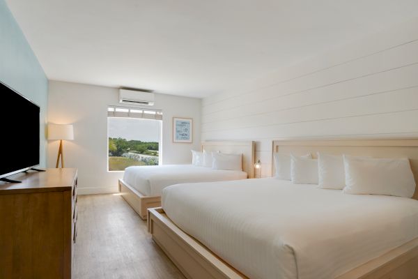 The image shows a clean, well-lit hotel room with two large beds, a window with a view, a TV, and a wooden dresser.