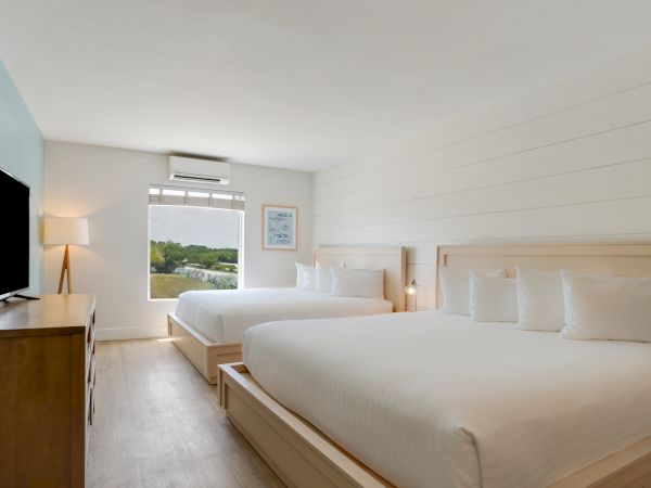The image shows a clean, well-lit hotel room with two large beds, a window with a view, a TV, and a wooden dresser.