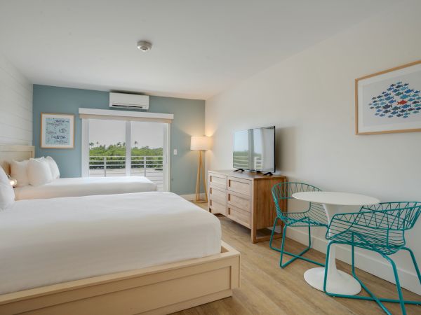 A modern hotel room with two beds, a TV, a small dining table with two chairs, and a large window with a view. The decor is minimalistic.