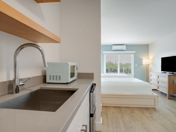 A modern studio apartment with a kitchenette featuring a sink and microwave, a cozy bedroom, a large window, and a flatscreen TV.