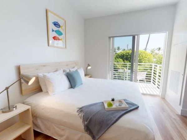 A bright bedroom with a large bed, bedside tables, a painting, a tray with items, a gray throw, and a balcony door leading outside.