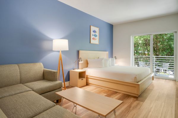 A modern, well-lit bedroom with a bed, sofa, lamp, coffee table, artwork on a blue wall, and a sliding door opening to a lush green view.