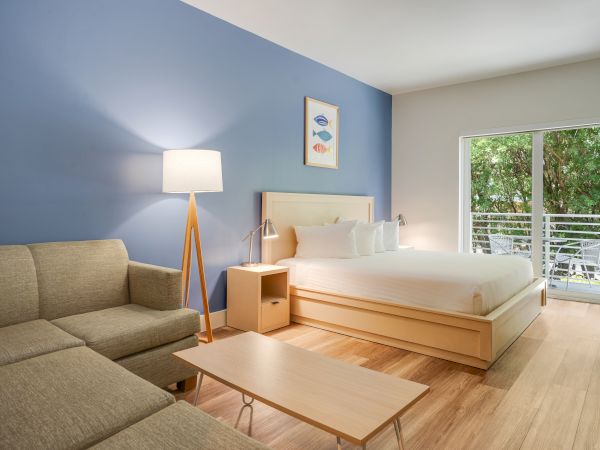 A modern, well-lit bedroom with a bed, sofa, lamp, coffee table, artwork on a blue wall, and a sliding door opening to a lush green view.