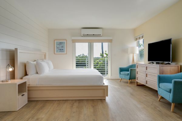 A modern, tidy bedroom with a large bed, two blue chairs, a flat-screen TV, wooden furniture, and a window with blinds letting in natural light.