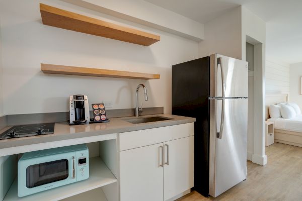 A small kitchen with a refrigerator, sink, coffee maker, microwave, two shelves, and an adjacent bedroom visible in the background.