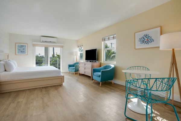 A modern, well-lit room with a bed, TV, two blue chairs, a dresser, and a small table with chairs on a wooden floor, and large windows.