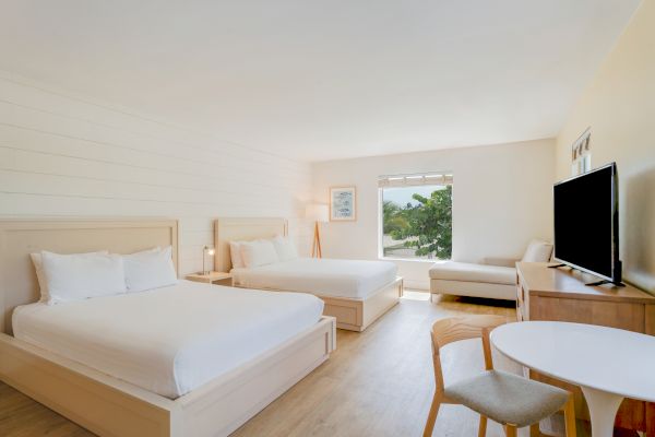 The image shows a hotel room with two beds, a TV, a small table and chairs, a window with a view, and minimalistic decor.