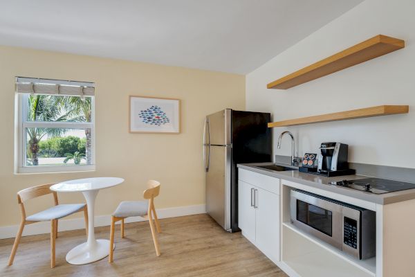 A modern kitchen area with a small dining table, two chairs, a window, a fridge, microwave, sink, coffee maker, and open shelving.