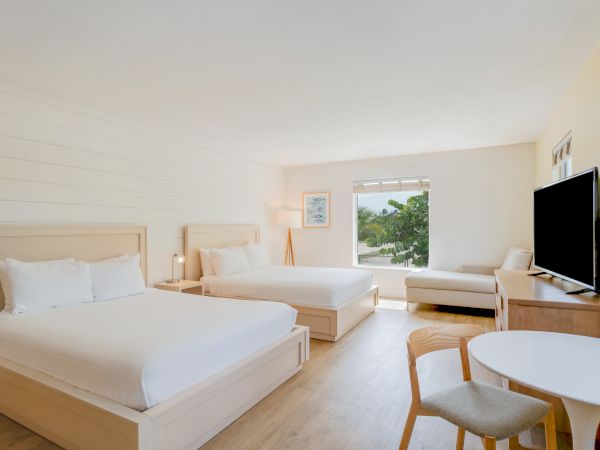 The image shows a hotel room with two beds, a TV, a small table and chairs, a window with a view, and minimalistic decor.