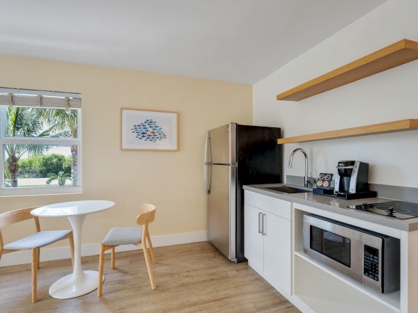 A modern kitchen area with a small dining table, two chairs, a window, a fridge, microwave, sink, coffee maker, and open shelving.