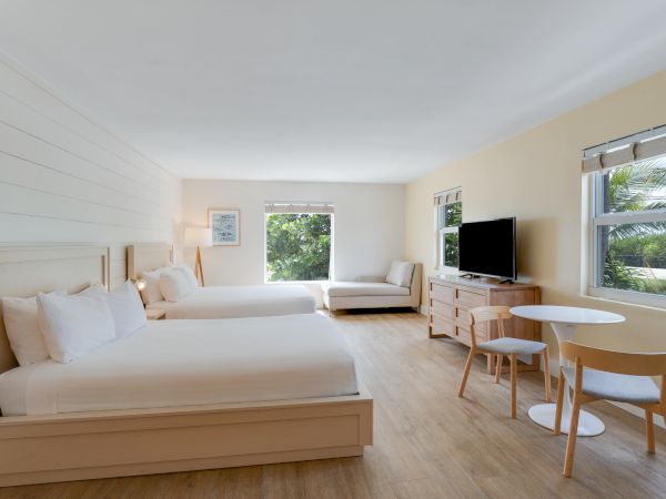 A spacious hotel room with two beds, a sofa, a TV, a round table with chairs, and windows allowing natural light in a bright, minimalist setting.