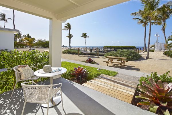 A beachside porch with two chairs and a small table, overlooking a pathway, picnic table, palm trees, sand, and ocean under a blue sky.