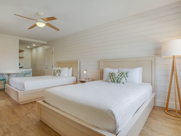 A room with two white beds, a floor lamp, a ceiling fan, and a small table with chairs, all set against a white wooden wall paneling.