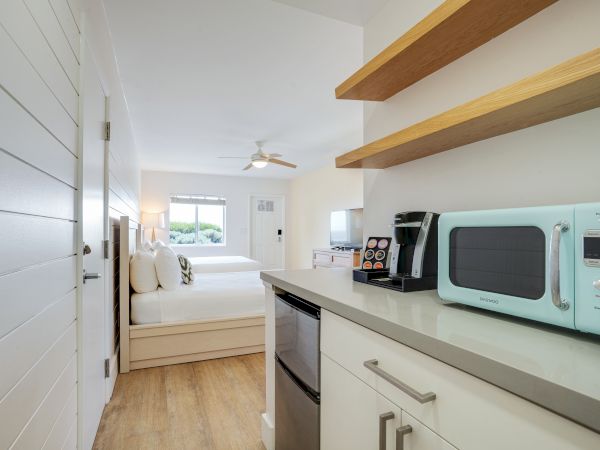 A modern hotel room features a kitchenette with a microwave, coffee maker, and mini-fridge, leading into a bedroom with two neatly made beds.
