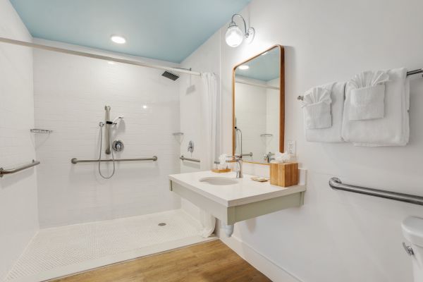 An accessible bathroom with a walk-in shower, grab bars, a floating sink, a mirror above, towels folded on a rack, and a wooden floor.