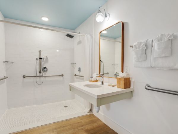 An accessible bathroom with a walk-in shower, grab bars, a floating sink, a mirror above, towels folded on a rack, and a wooden floor.