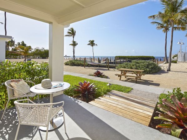 A cozy patio with a table and two chairs overlooks a beach area with palm trees, lush plants, and a picnic table facing the ocean.