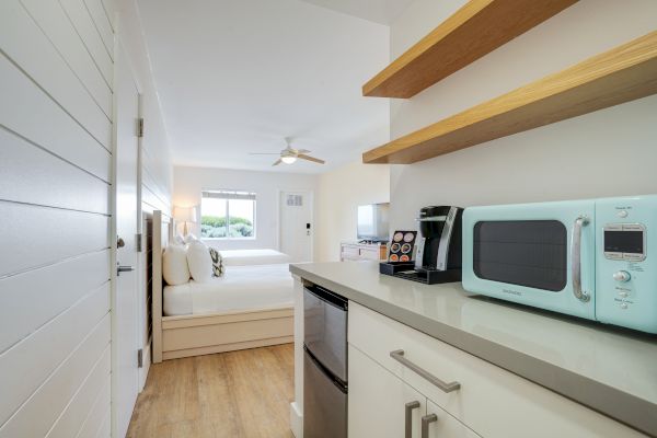 A modern hotel room with a kitchenette featuring a microwave, coffee maker, and shelves. The room has two beds, a ceiling fan, and a window.