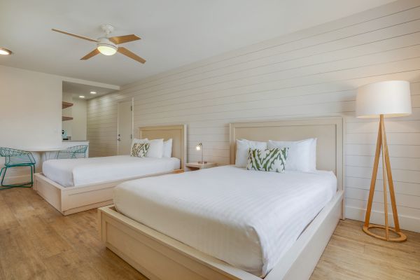 A bedroom with two double beds, a floor lamp, a ceiling fan, wooden flooring, and a small dining table with chairs in the background.