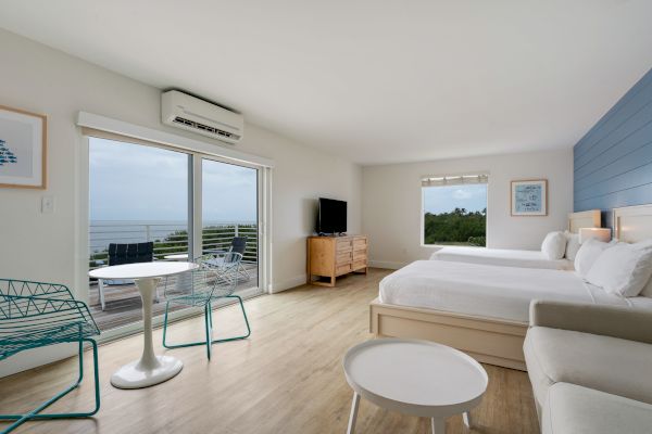 A spacious hotel room features two beds, a TV, a sofa, and a table with chairs, with a sliding door leading to a balcony overlooking the sea.