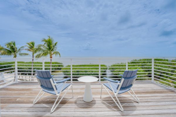 Two chairs and a small table on a wooden deck overlook a tropical seascape with palm trees, greenery, and a cloudy sky in the background.