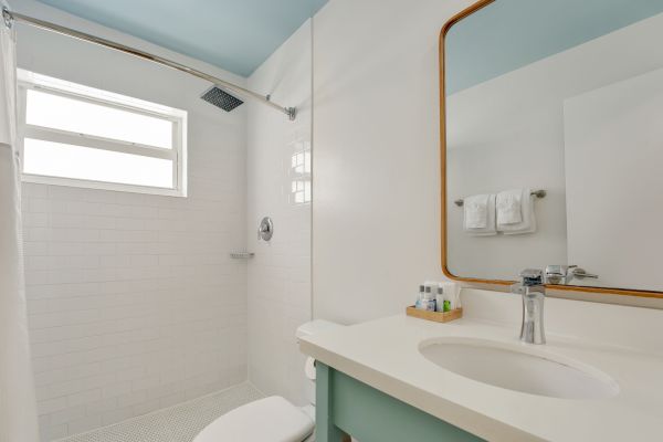 This image shows a clean bathroom with a shower area, a sink with a faucet, a large mirror, toiletries on the counter, and a window.