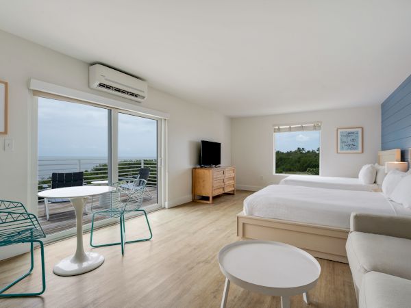 A spacious hotel room features two beds, a TV, a sofa, and a table with chairs, with a sliding door leading to a balcony overlooking the sea.