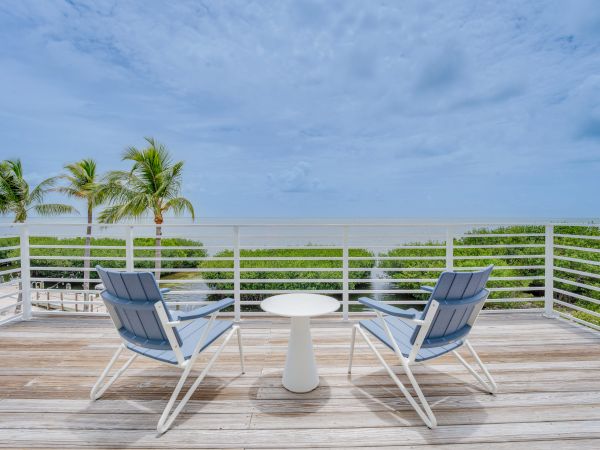 Two chairs and a small table on a wooden deck overlook a tropical seascape with palm trees, greenery, and a cloudy sky in the background.