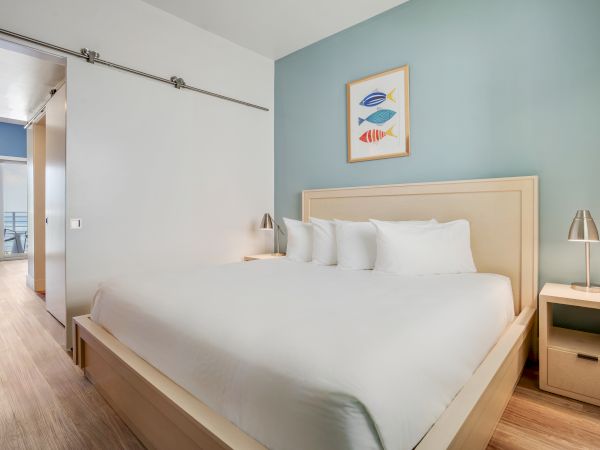 A modern bedroom features a large bed, two side tables with lamps, and a fish-themed picture. The room extends to a balcony area.