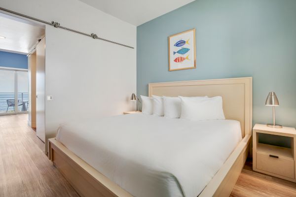 The image shows a modern bedroom with a large bed, three pillows, two bedside tables with lamps, and a picture of fish above the bed.