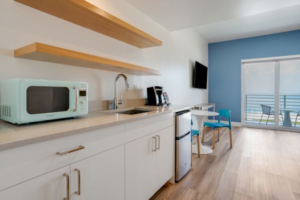 A modern kitchenette with white cabinets, a blue microwave, a coffee maker, a mini-fridge, a TV, and a table with chairs near a balcony.