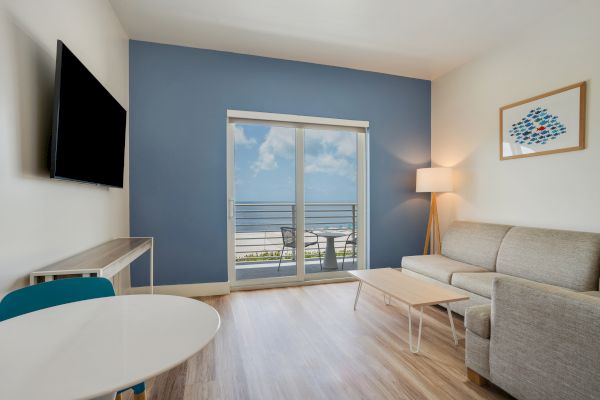 This image shows a modern living room with light wood flooring, a sofa, coffee table, lamp, wall art, TV, and a view of the ocean through a glass door.