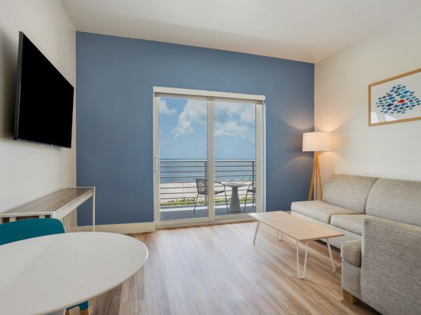 This image shows a modern living room with light wood flooring, a sofa, coffee table, lamp, wall art, TV, and a view of the ocean through a glass door.