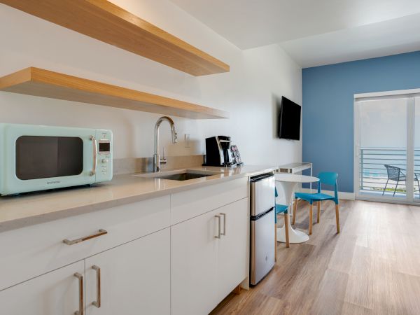 A modern kitchenette with white cabinets, a blue microwave, a coffee maker, a mini-fridge, a TV, and a table with chairs near a balcony.