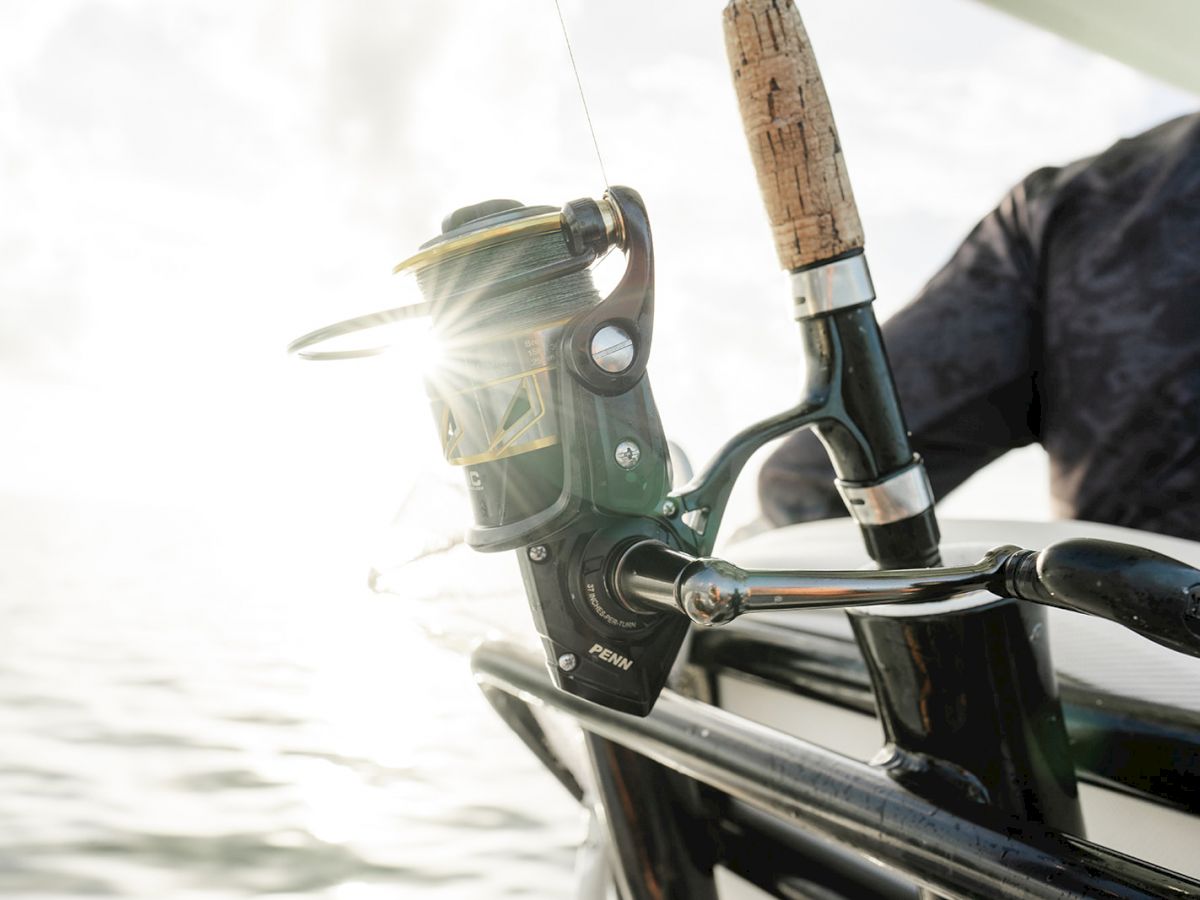 The image shows a close-up of a fishing rod and reel held by a person on a boat, with sunlight glinting off the reel.