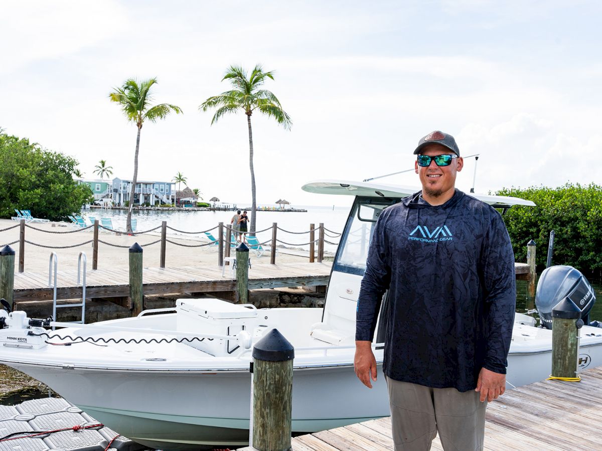 A man wearing sunglasses and a long-sleeve shirt stands on a dock next to a white boat, with palm trees and water in the background.