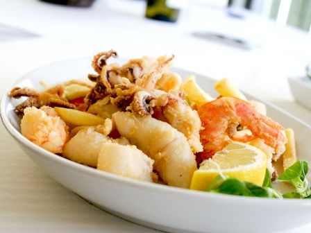 The image shows a bowl of mixed fried seafood including shrimp, calamari, and fish, garnished with lemon and green herbs on the side.