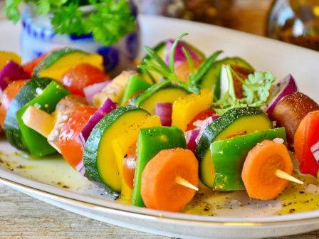 The image shows colorful vegetable kebabs on skewers, including carrots, bell peppers, zucchini, onions, and tomatoes, placed on a white plate.