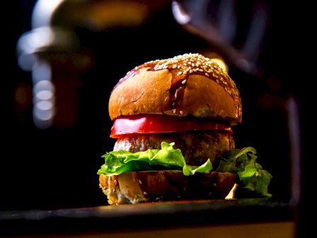 A close-up image of a hamburger with lettuce, tomato, and a sesame seed bun is seen against a dark background, highlighting its juicy and fresh appearance.