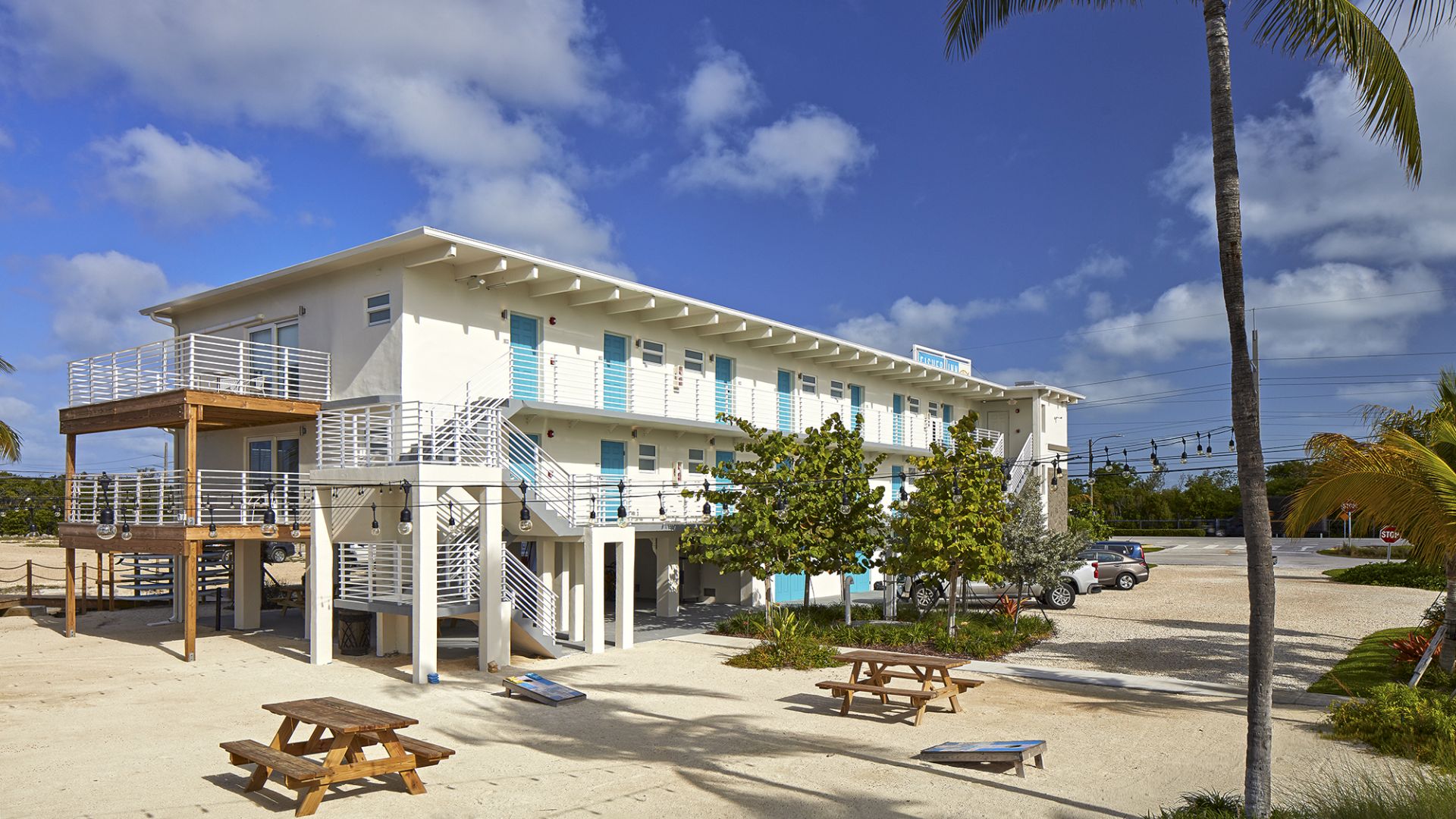A modern, multi-story building with turquoise shutters, picnic tables, and palm trees in the foreground under a bright blue sky with scattered clouds.