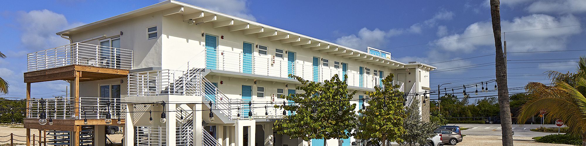 A modern, multi-story building with turquoise shutters, picnic tables, and palm trees in the foreground under a bright blue sky with scattered clouds.