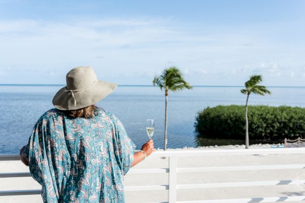 A person wearing a hat and holding a drink enjoys a scenic ocean view from a balcony or deck with palm trees and a clear sky.