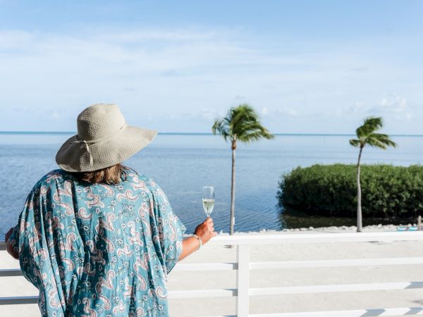A person wearing a hat and holding a drink enjoys a scenic ocean view from a balcony or deck with palm trees and a clear sky.