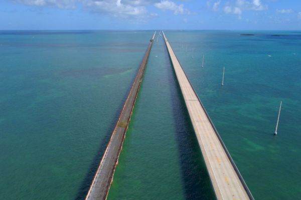 The image shows two parallel bridges stretching over a turquoise ocean, leading towards a distant horizon under a partly cloudy sky.