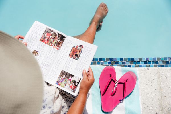 A person sits poolside reading a magazine, with legs dipped in the water, next to a striped towel and a pair of pink flip-flops.